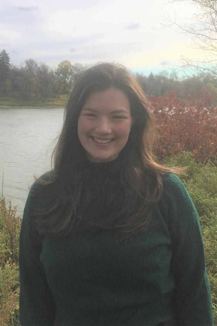Outdoor portrait of Lindsay Inglis standing near a river, surrounded by fall foliage under an overcast sky.