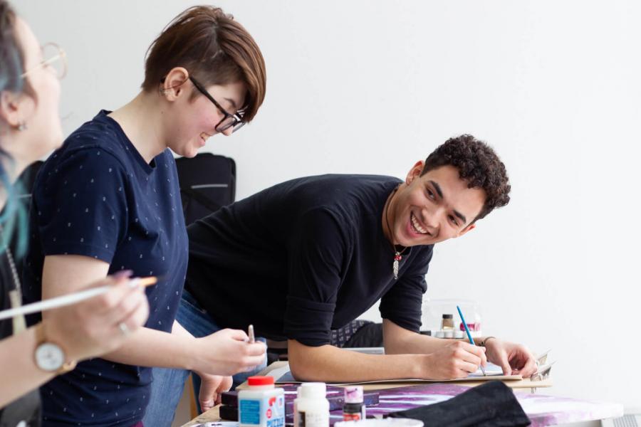 Three students stand at a narrow bench working on painting projects.