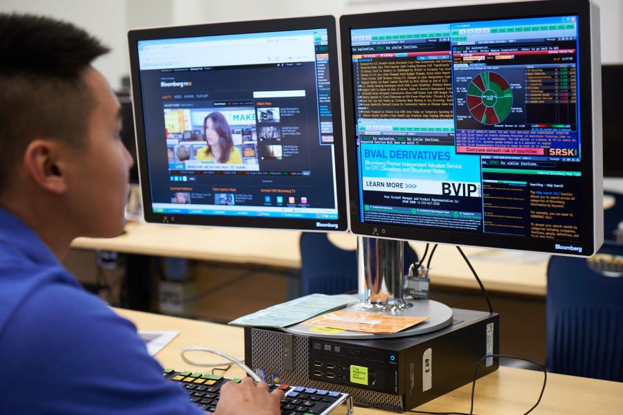 A student sits in front of a computer with two monitors with various programs running while working.