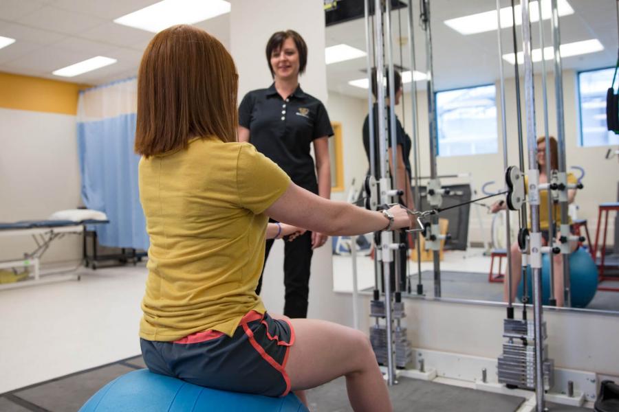A student works in the athletic therapy lab with a person sitting on a ball working with a piece of equipment.