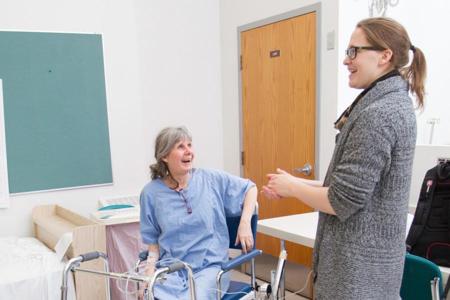 A nursing student laughs while talking to a senior who is sitting in a wheelchair nearby.