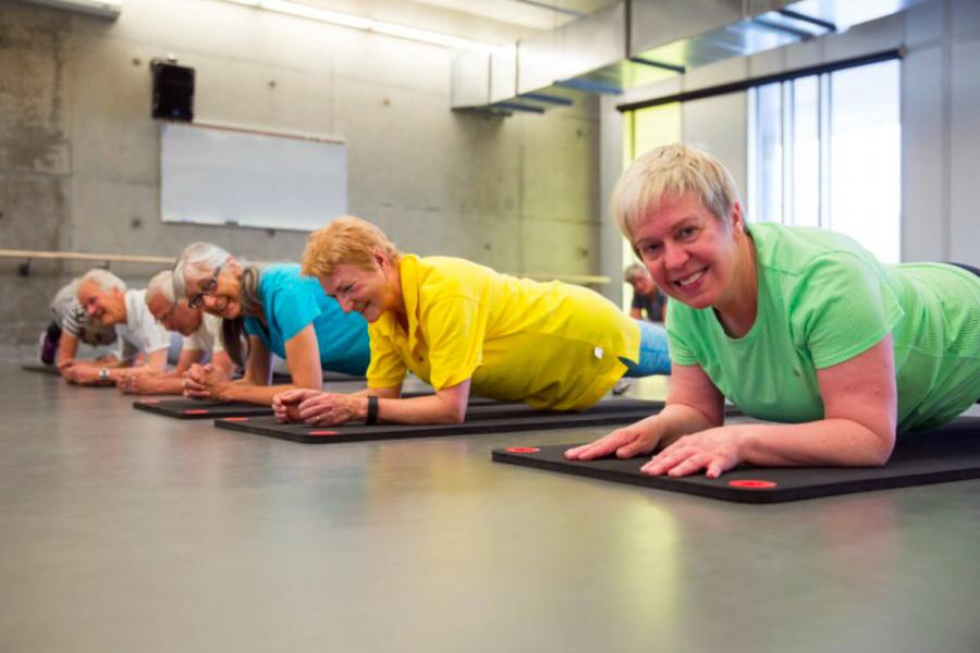 Six smiling seniors balance on their elbows and toes performing plank exercises on black mats.