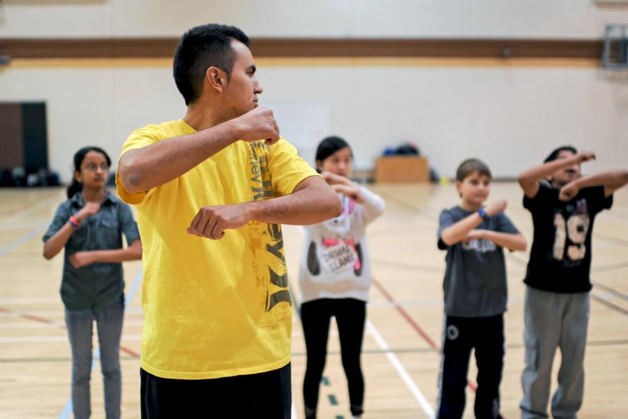 A physical education student teacher demonstrates a warm up skill to a class of young children standing in a row in a gymnasium.