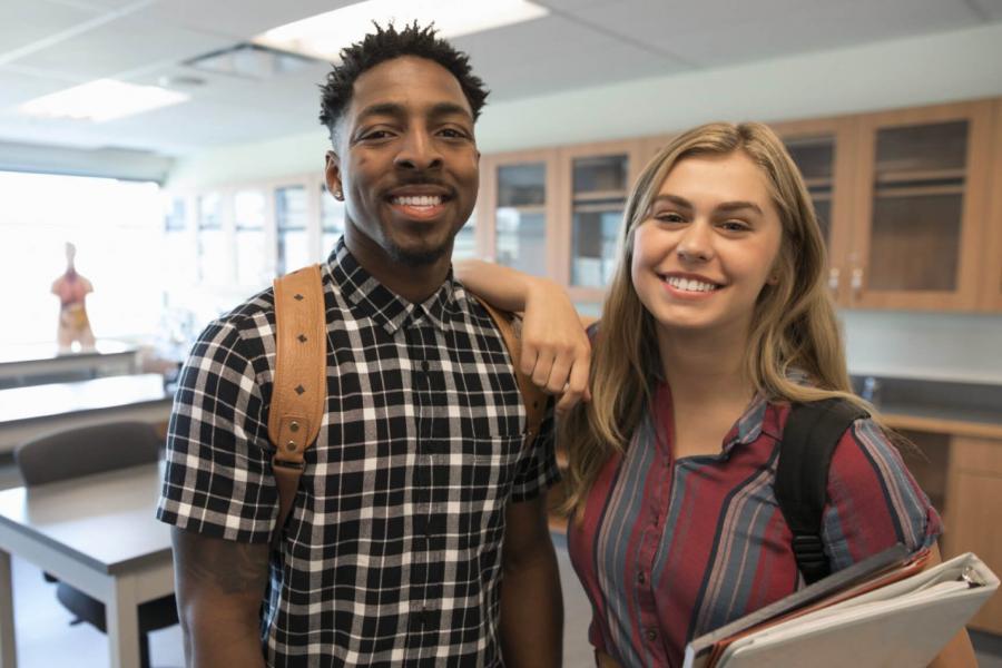 Two smiling recreation management students stand side by side holding books in a classroom. 