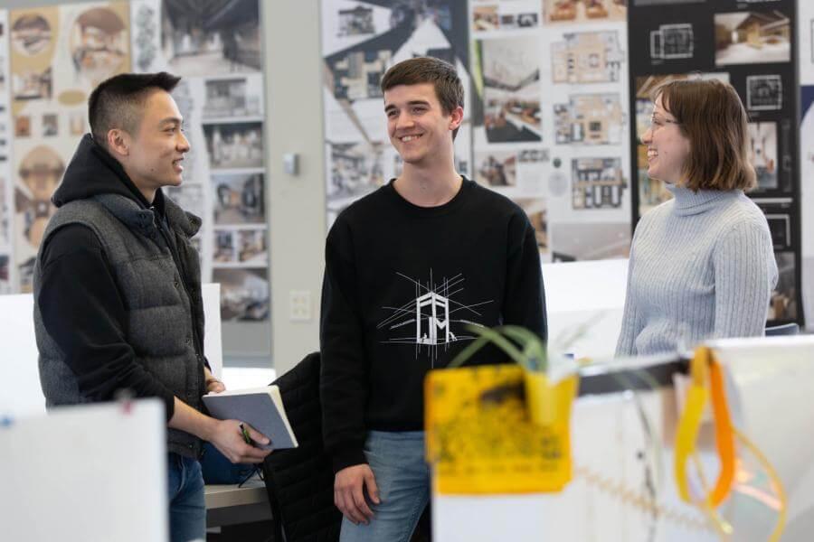 Three students stand together talking at their desks. 