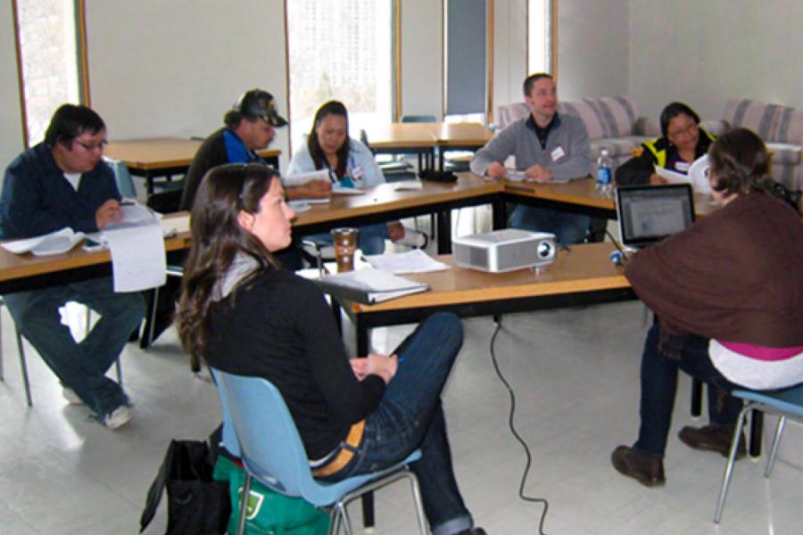 Indigenous students sit at three tables that form a triangle and listen to an instructor.