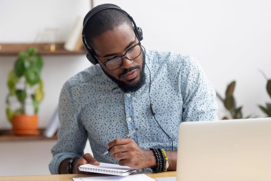 Man looking down at laptop, writing something on notepad.