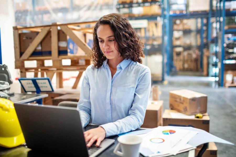 A woman stands at a desk in a warehouse and types on a laptop.