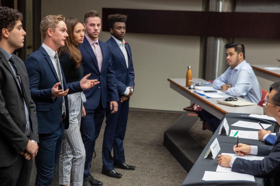 Five students stand at the front of a room giving a presentation to a panel of people.