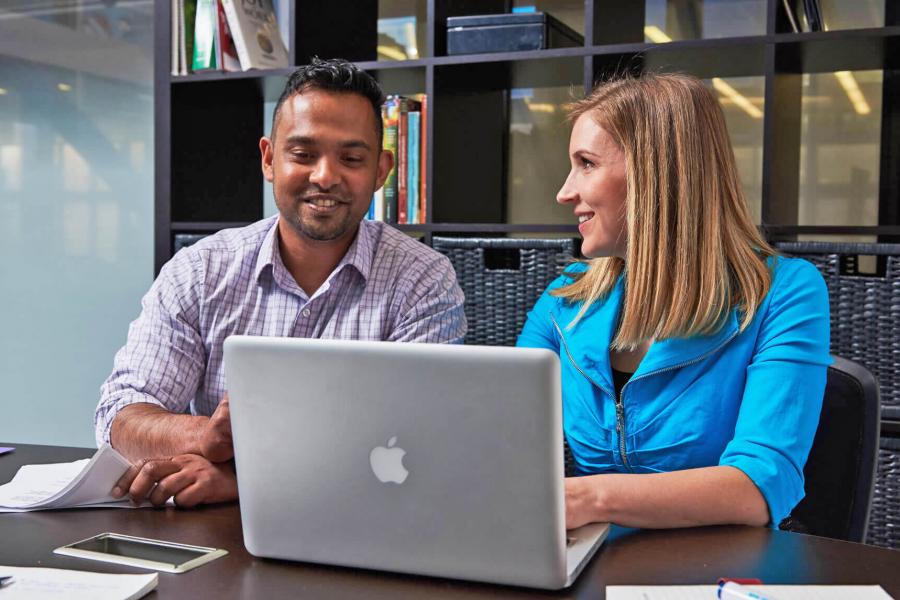 Two people sit together at a desk and look at a laptop monitor. 