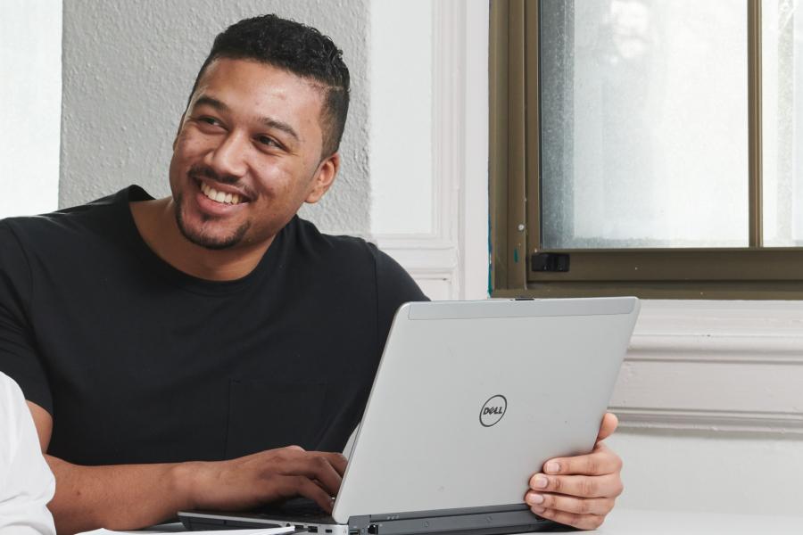 A student smiles while seated at a desk working on a laptop.