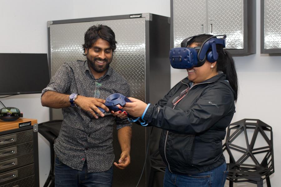 An engineering student assists another using a virtual reality headset and controller.