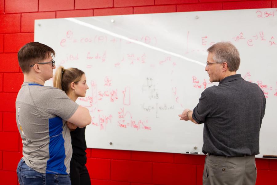 Two engineering students stand at a white board and listen to a professor.