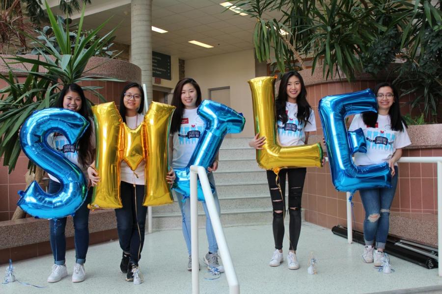 Five students stand side by side holding balloons which spell out the word smile.