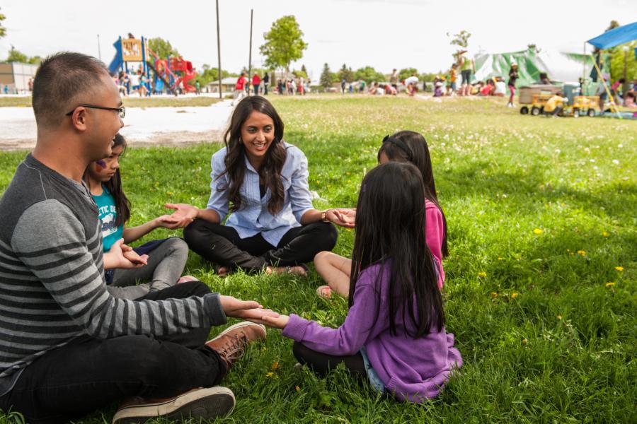 Two teachers sit with several students cross-legged in the grass forming a circle.