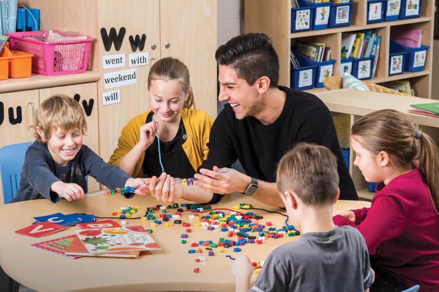 A smiling teacher sits at a table with four young students.