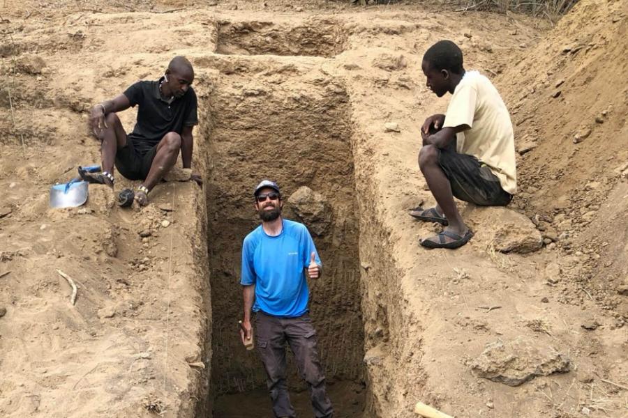 A geological scientists stands with two coworkers at a dig site.