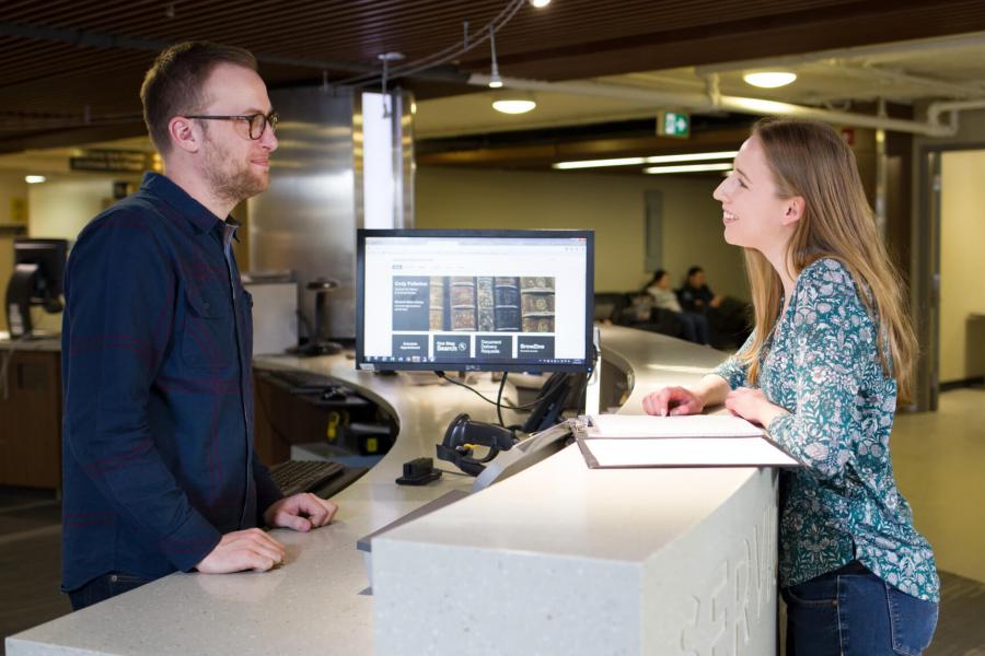 An arts student speaks with someone at a desk in a library.