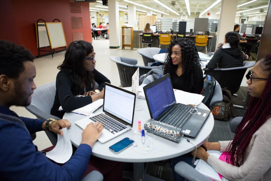 Students seated at a table using laptops and talking.