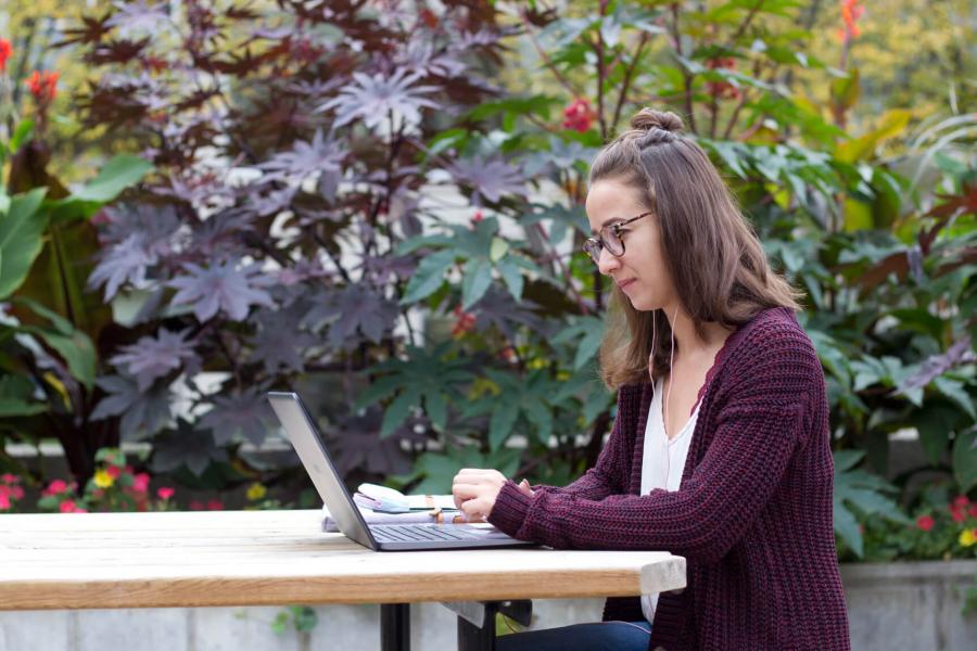 A student types on a laptop while seated at a table in a well landscaped outdoor area.