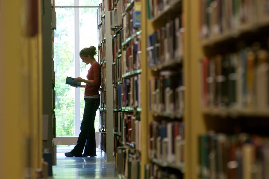 A student leans against a shelf of books while reading. 