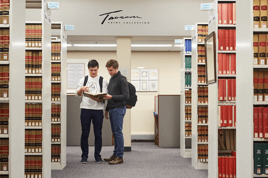 Two students look at a book together in a library. 