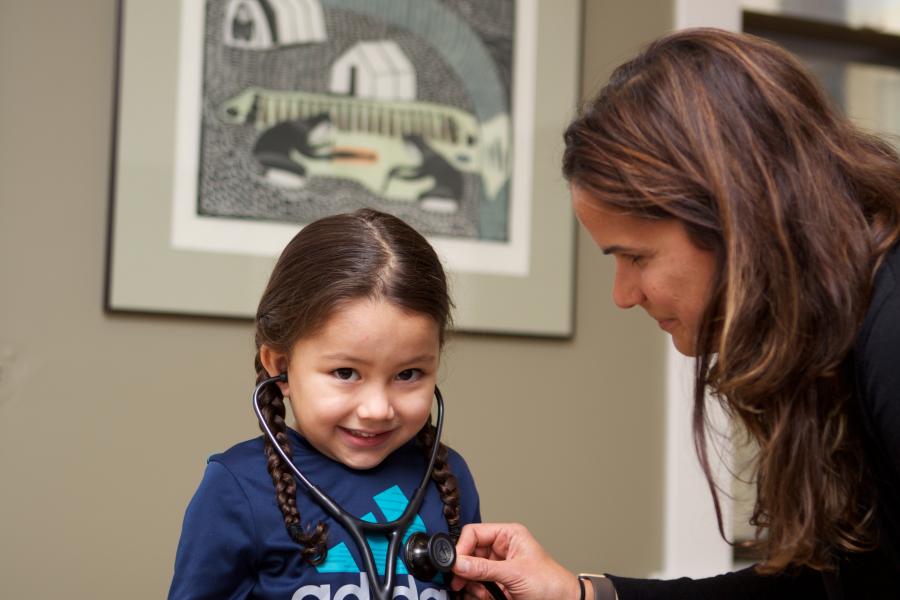 A small, smiling child is seated on a table with a stethoscope in their ears. A student holds the stethoscope to the child's chest so they can hear their heartbeat.