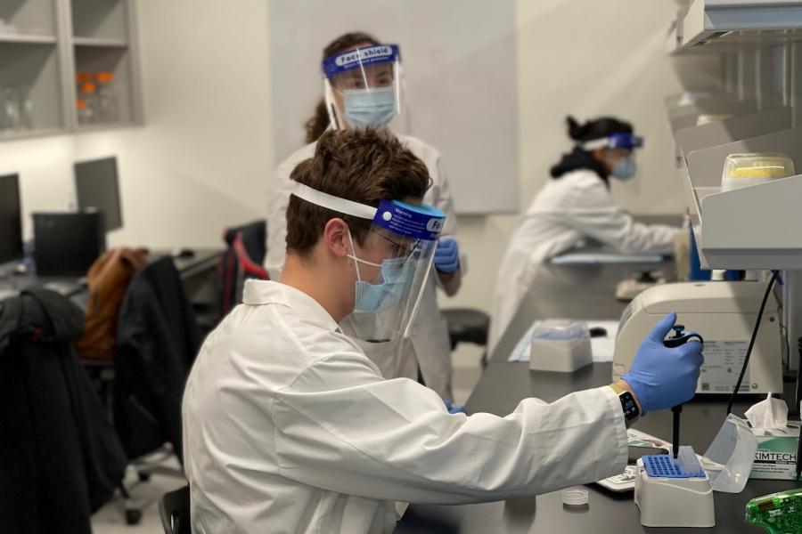 A student in personal protective equipment (PPE) is using a liquid dropper to collect samples in a lab. Two other students in PPE are visible in the background.