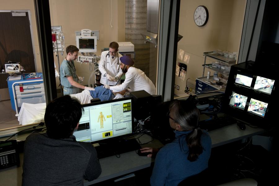 Wide shot of three medicine students performing an examination on a dummy, with one student holding a stethoscope to its chest. Two instructors observe the students from an observation room on the other side of a pane of glass.