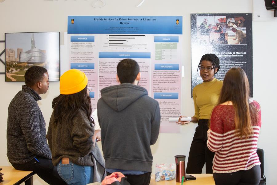 A woman gives a presentation to a group of four students. A poster in the background reads "Health Services for Prison Inmates: A Literature Review."