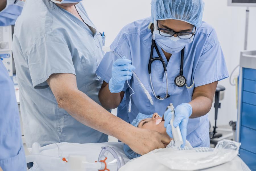 A student in wearing blue scrubs and personal protective equipment shines a light into the mouth of a patient on an examination table. A surgeon supervises the student while another student stands slightly out of frame. 