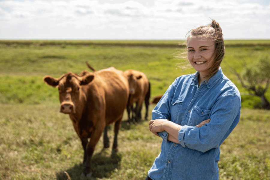 a smiling student stands in a field with two cows standing in the background.
