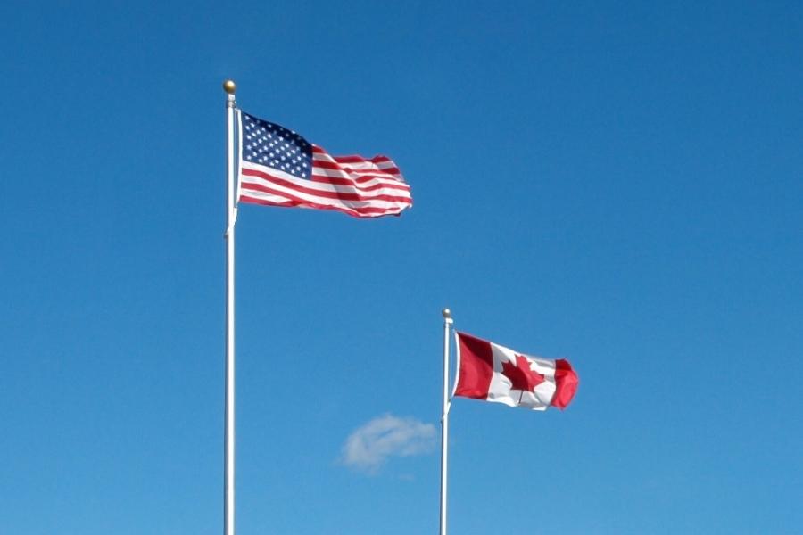 A United States of America and Canadian flag fly side by side against a blue sky.