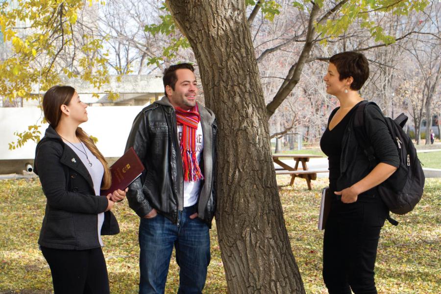 Three Indigenous students stand together having a conversation outdoors next to a tree.
