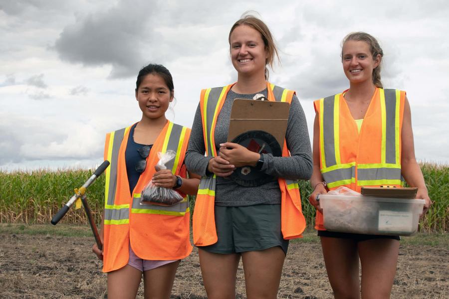 Three students in high visibility vest stand in a field holding soil sampling equipment.