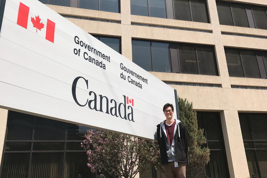 A student stands in front of a building and a sign that says Government of Canada.