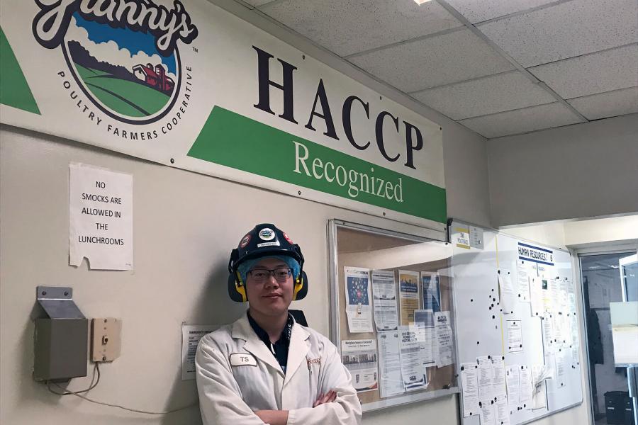 A coop student wearing a lab coat and hard hat stands in front of a sign.