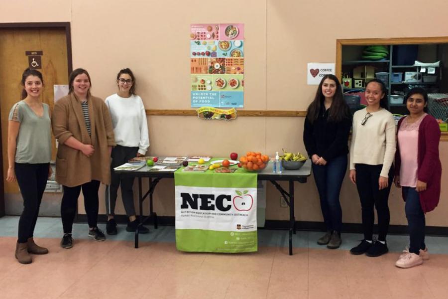 A group of students stands by a table with fruit and a sign that says NECO.