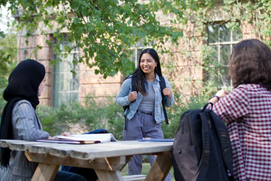 Three students say hello to each other as they walk outdoors. Two are seated at a picnic table. 