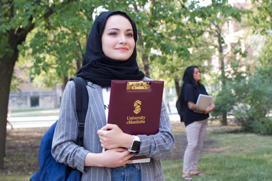 A student stands smiling, her arms crossed in front of her as she holds a clipboard with the University of Manitoba logo on it. 