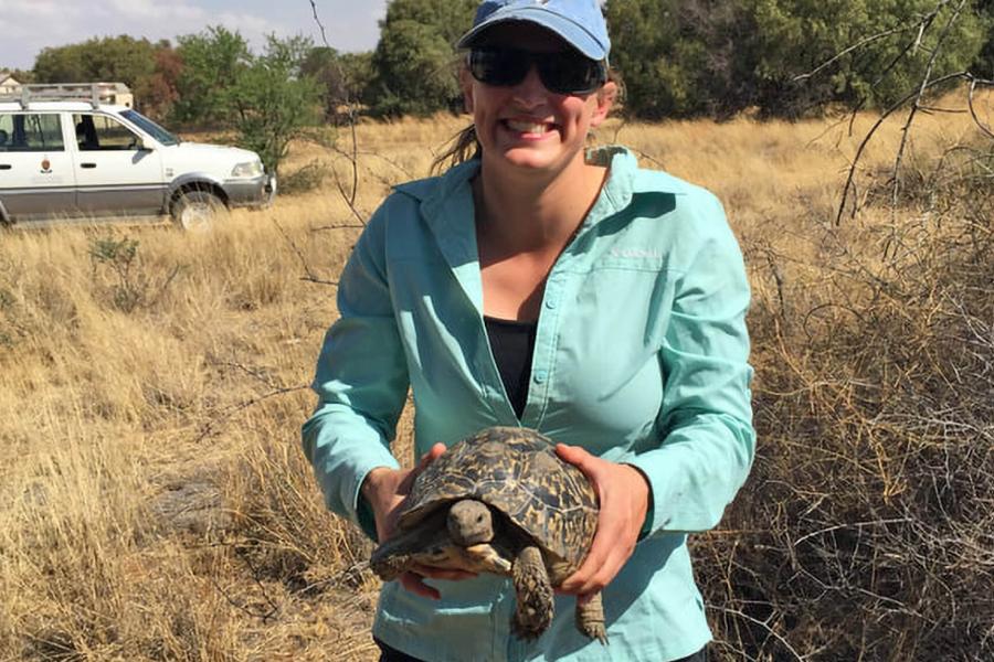 Krista Shofstall a biological sciences PHd student stands in a field smiling while holding a turtle with both hands in front of her.