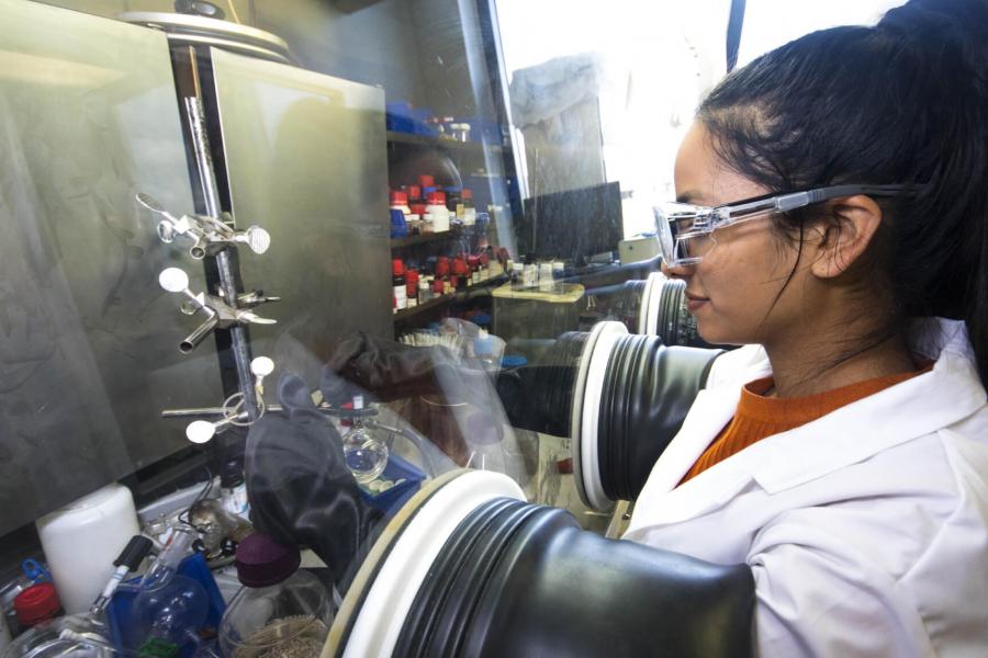 A student wearing gloves, glasses and a white coat works carefully with equipment in a lab.