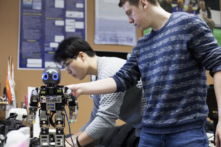 Two male students work on a small robot with bright blue circular eyes. 