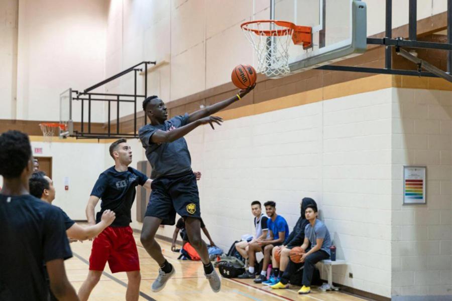 Several students look on from a bench as others play recreational basketball in a gym. One player is in mid air under the net attempting to score.