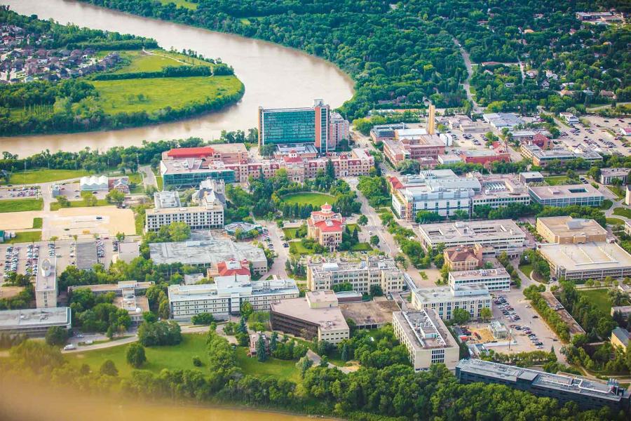 Aerial view of the Fort Garry Campus. More than sixty traditional and modern buildings are surrounded by lush trees and the curve of a river.
