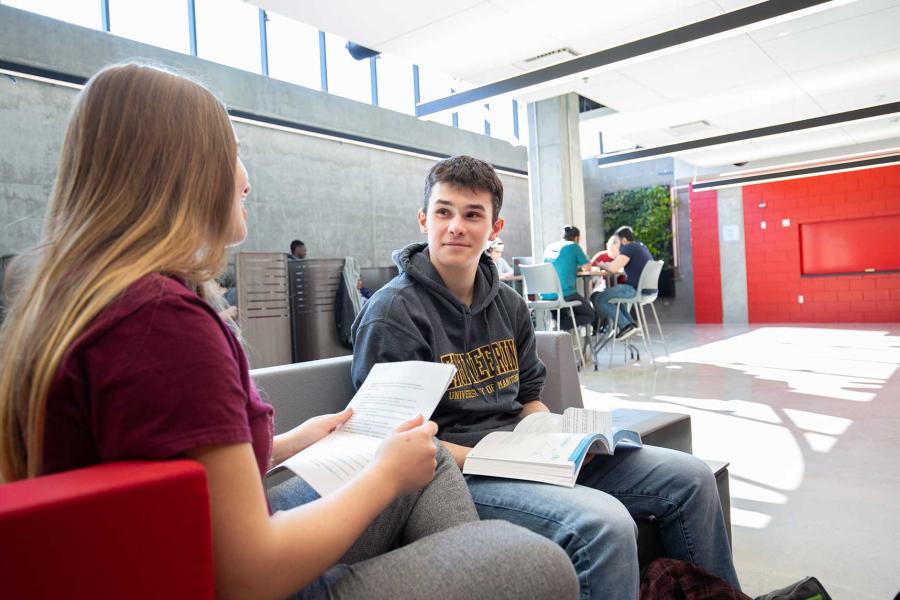 Two students sitting on plush chairs and holding textbook materials in a large student lounge. 