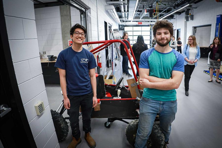 Two smiling students in a workshop posing in front of a small four-wheeled vehicle they have designed and built.