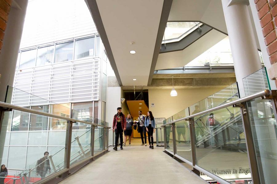 Students walk along a walkway with glass and metal railings along each side. 