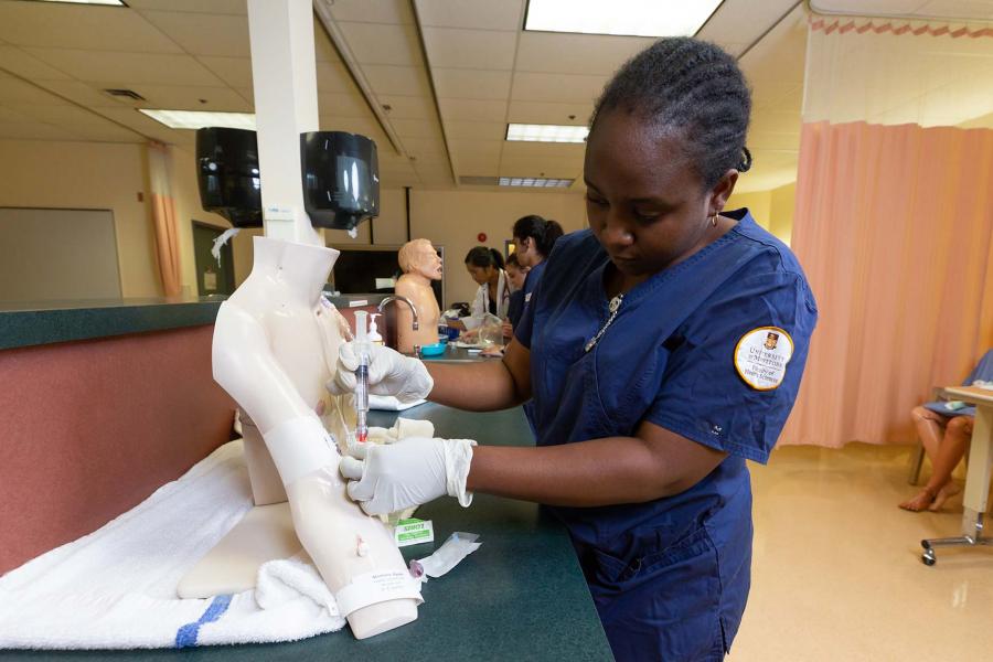 A woman in scrubs stands at a counter inserting an IV into a plastic arm.