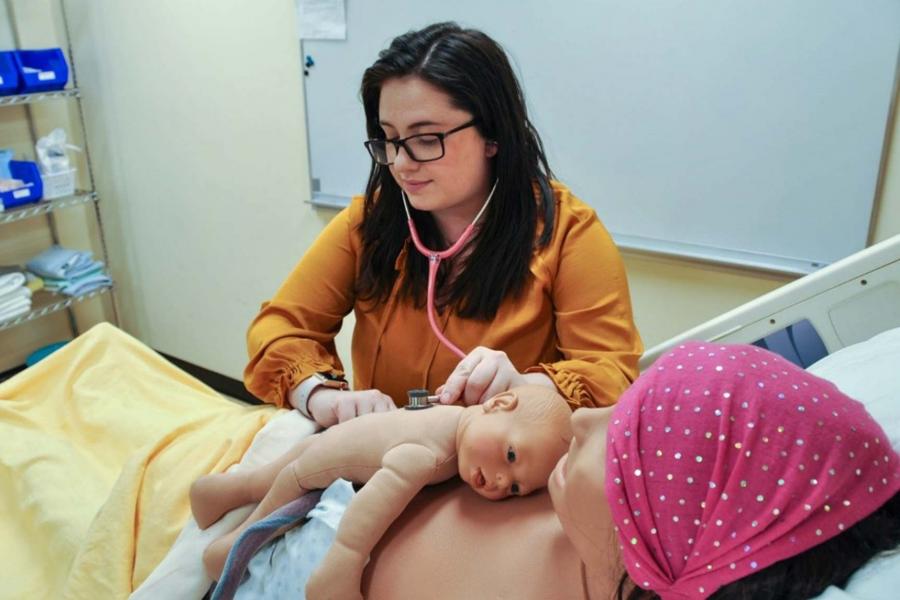 A student checks the heartbeat of an infant mannequin lying on the chest of an adult mannequin in a hospital bed.